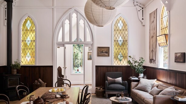 A cozy room with a wooden table, a sofa, and a fireplace. There are large windows with stained glass, a chandelier, and a door leading outside. - The Church at Lyonville ~ Daylesford Region (Lyonville)