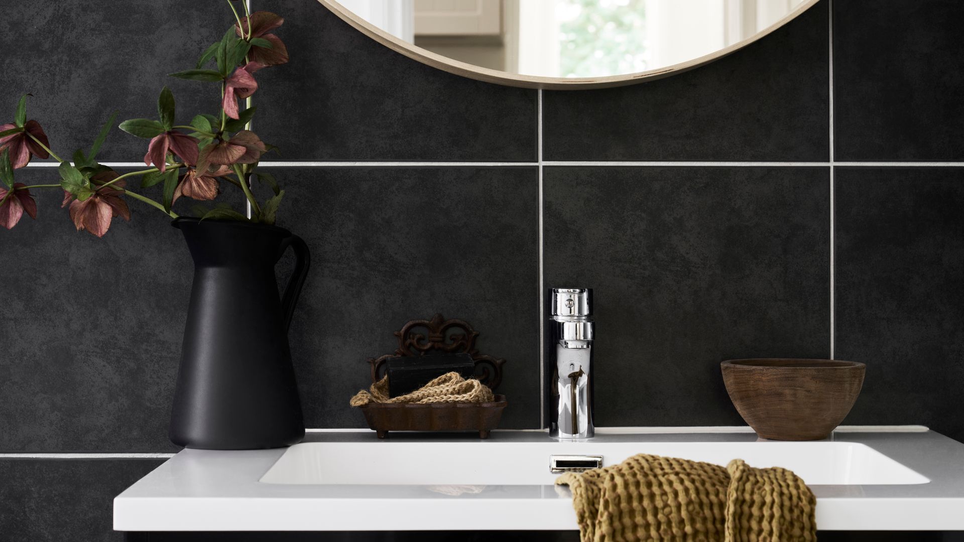 Une salle de bain avec un lavabo blanc, un miroir rond, un vase noir avec des fleurs, un petit bol en bois et une serviette tissée.
