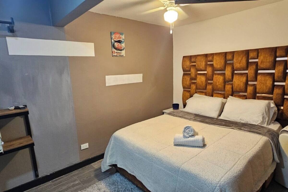 A hotel room with a bed, a small table with a microwave, a wall-mounted shelf, and a ceiling fan.