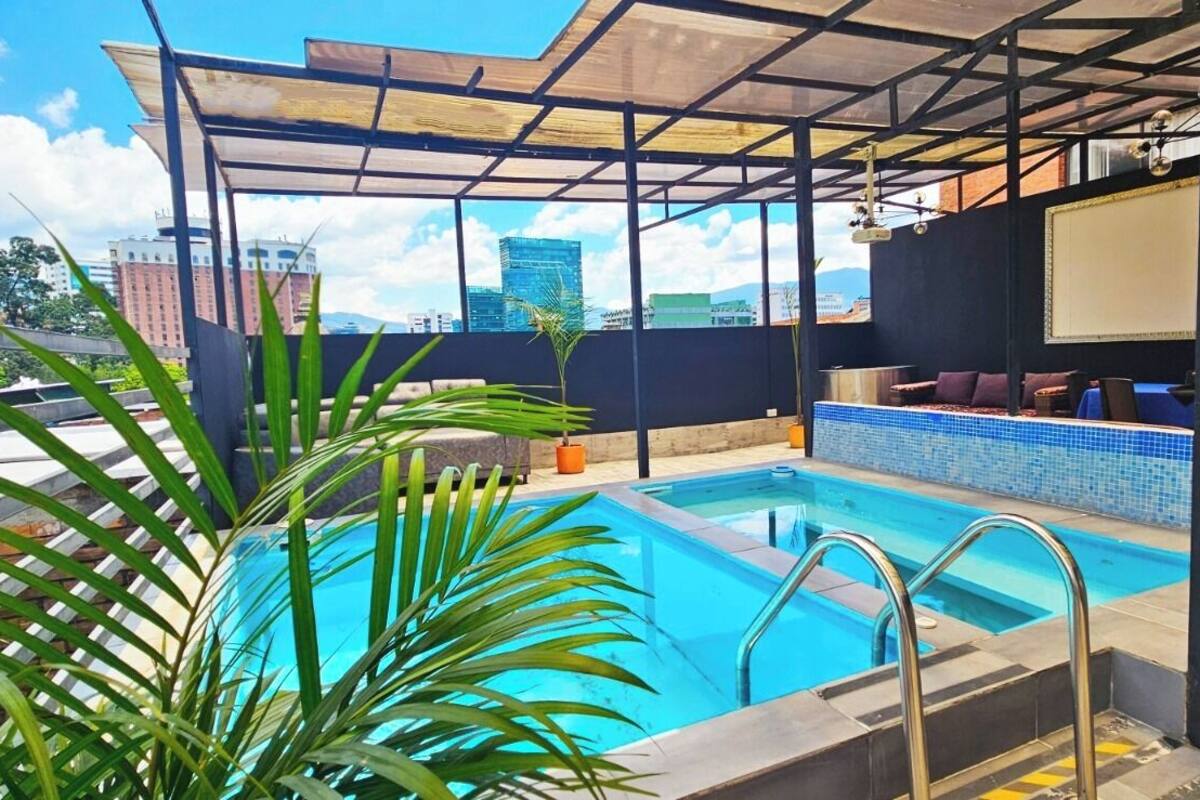 A rooftop pool with a view of city buildings, a covered seating area, and potted plants.