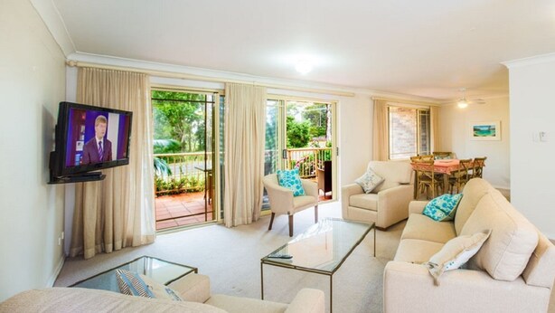 A living room with a sofa, armchairs, a glass coffee table, a TV, and a dining area with a table and chairs. - Fully furnished 2 bedroom apartment (Mollymook)