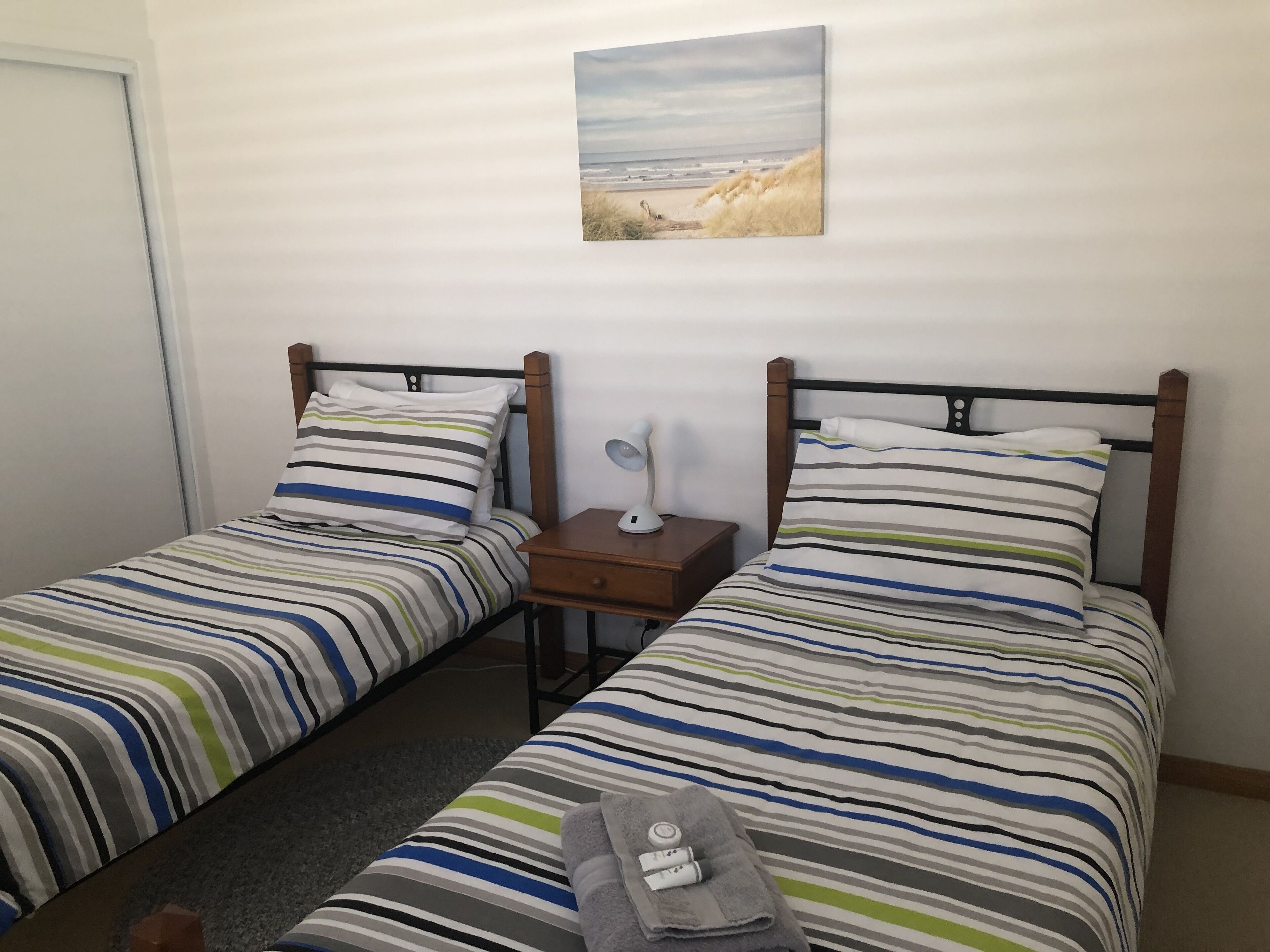 Ein Zimmer mit zwei Einzelbetten, jeweils mit gestreifter Bettwäsche, einem Nachttisch mit Lampe sowie einem Bild einer Strandlandschaft an der Wand.