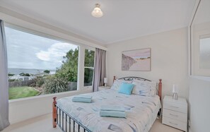 A bedroom with a large window, a bed with blue bedding, a nightstand, and a wall-mounted lamp. - Fabulous Ocean Views (Bridport)