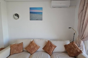 A living room with a sofa, decorative pillows, a wall clock, a painting of a beach, and an air conditioning unit.