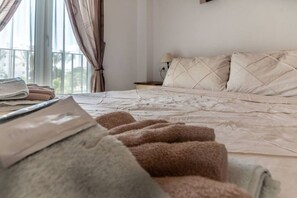 Una cama bien hecha con cabecera acolchada, lámpara de noche y ventana con cortinas.