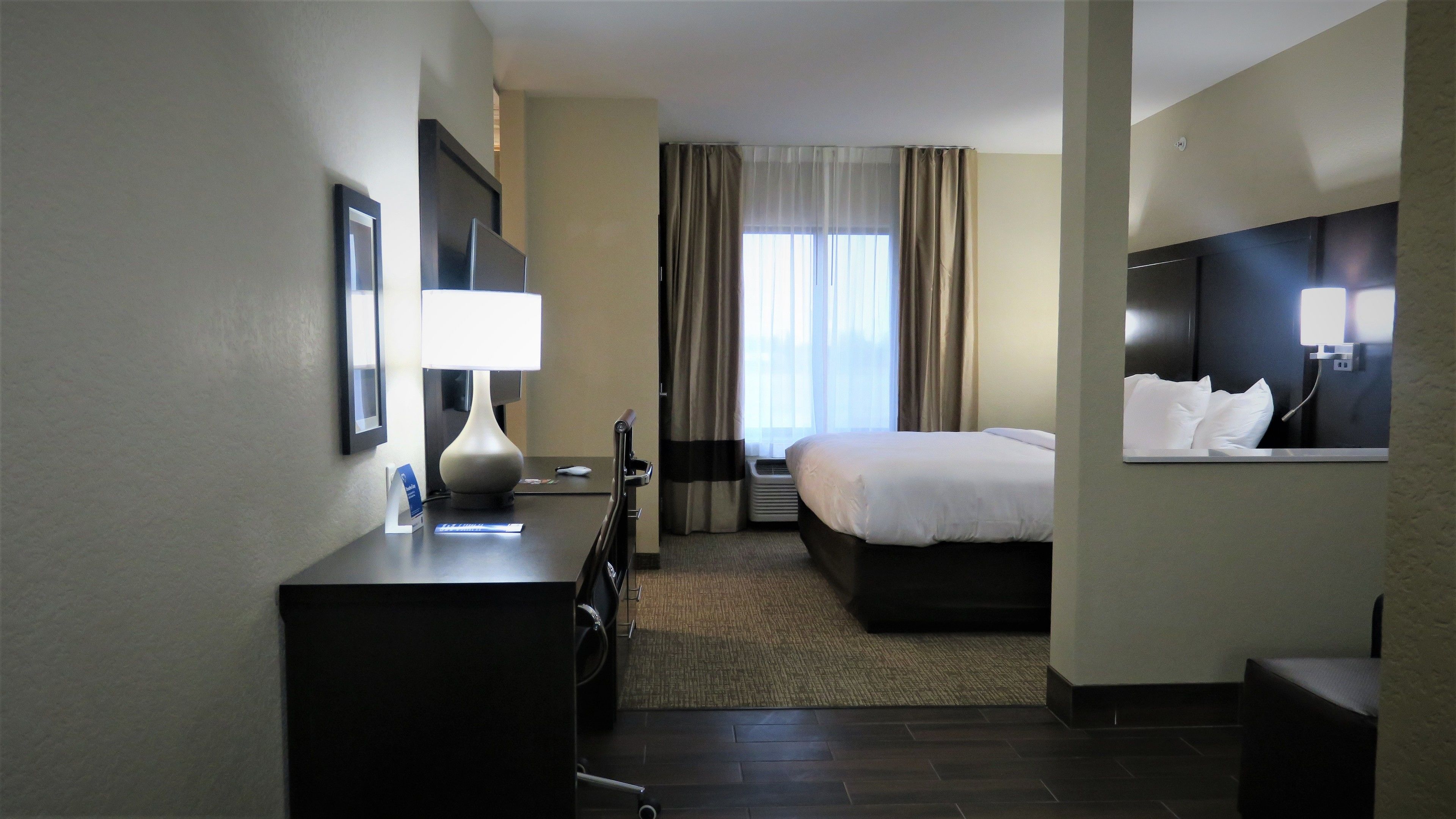 Une chambre d’hôtel comprenant un lit, un bureau avec une lampe, une chaise, une fenêtre avec des rideaux et un téléviseur fixé au mur.