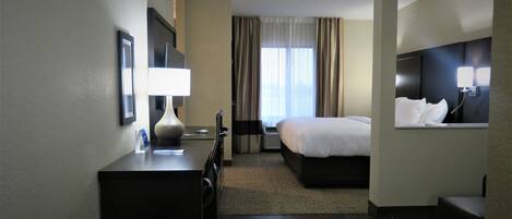 A hotel room with a bed, a desk with a lamp, a chair, a window with curtains, and a wall-mounted TV.