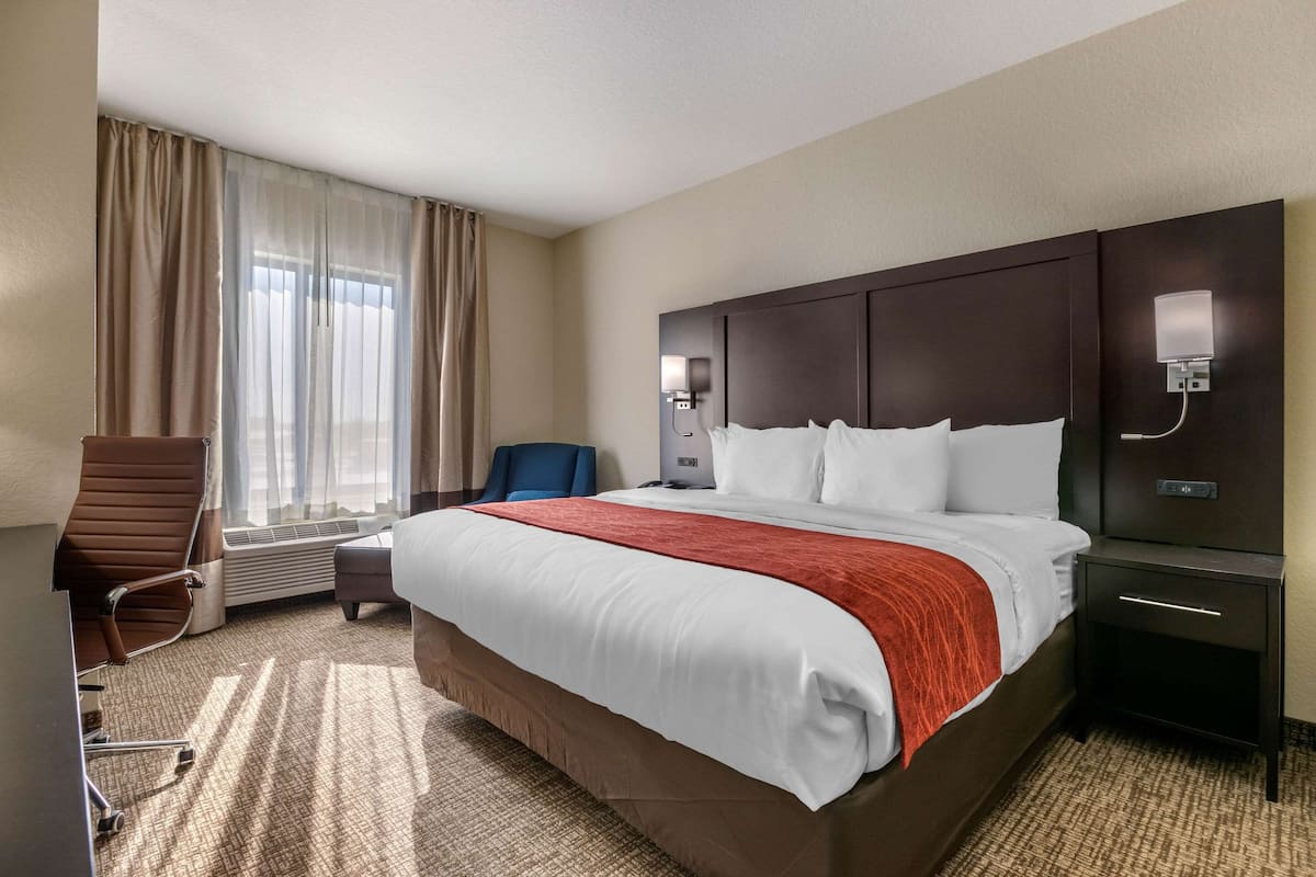 Habitación de hotel con una cama grande, un escritorio, una silla, un ventanal con cortinas y una mesita de noche con lámpara.