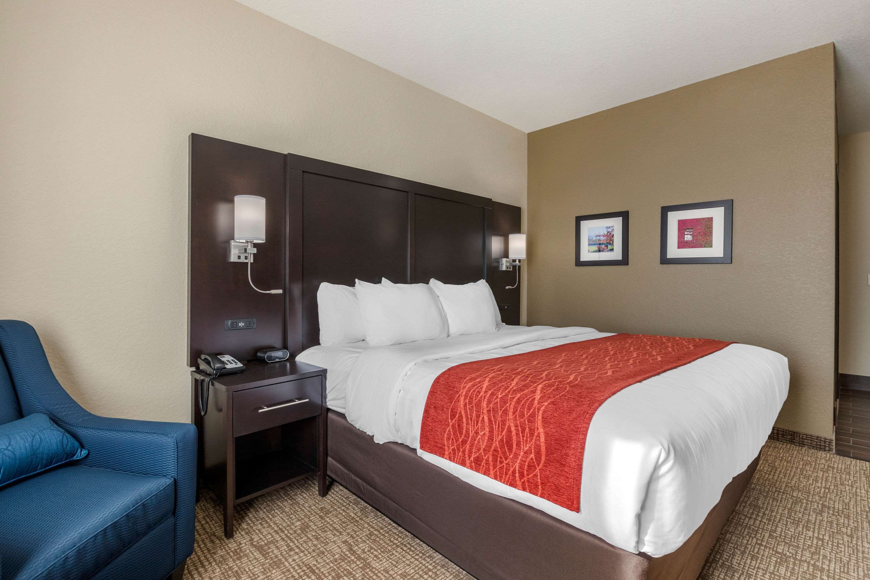 A hotel room with a large bed, a bedside table with a phone, a blue armchair, and two framed pictures on the wall.
