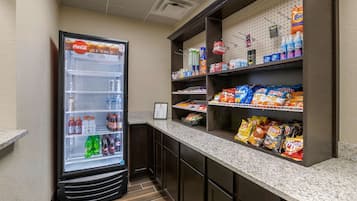 Un refrigerador de autoservicio con diversas bebidas, una barra de aperitivos con papas fritas y otros bocadillos, y un piso limpio con azulejos.
