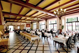Um amplo salão de jantar com tetos altos, lustres e um padrão de piso xadrez. Mesas montadas com toalhas brancas e cadeiras.