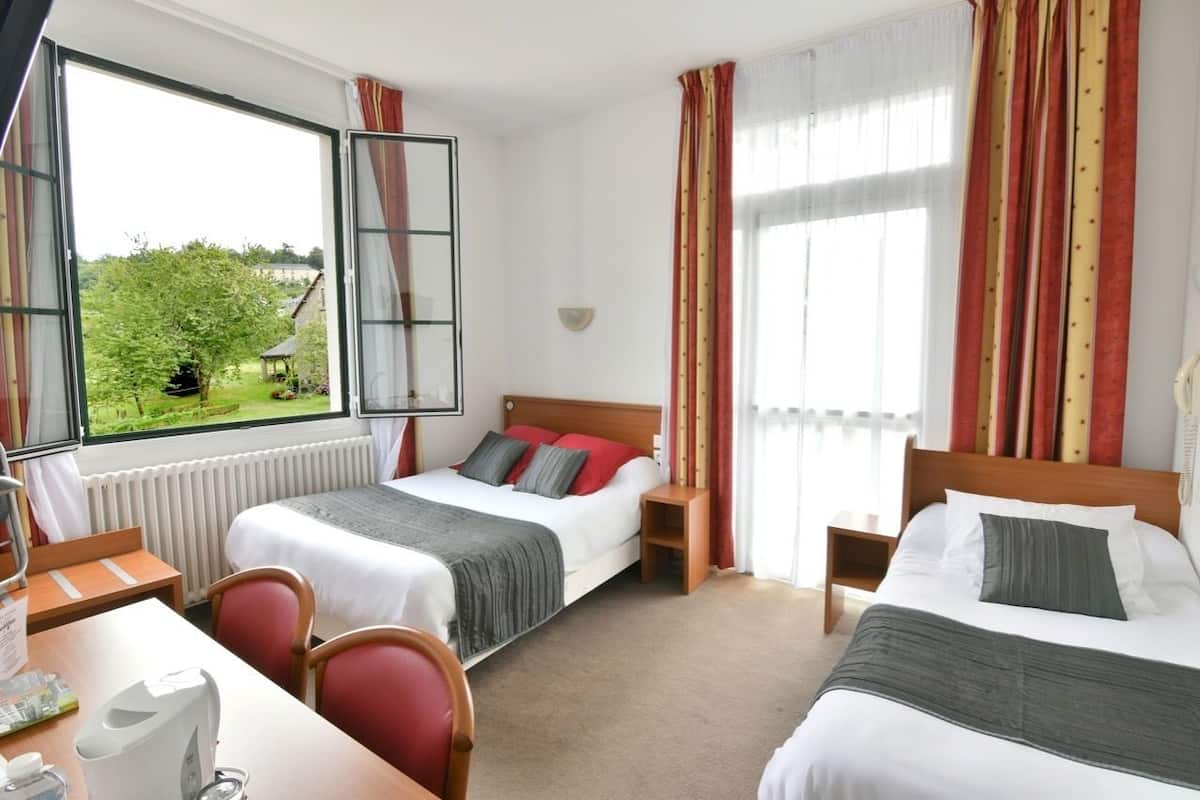 A hotel room with two beds, a desk, and a chair. There is a window with curtains, a radiator, and a view of greenery outside.
