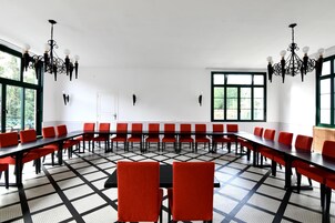Uma sala de conferências com mesas compridas e cadeiras vermelhas, janelas amplas e lustres pretos.