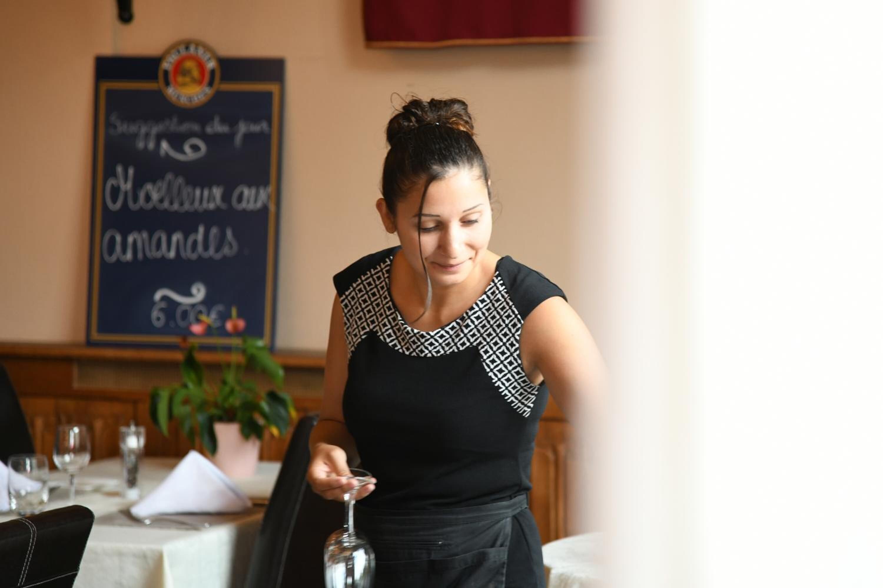 Eine Person serviert ein Glas Wein an einem Restauranttisch, im Hintergrund ein Kreidetafel.