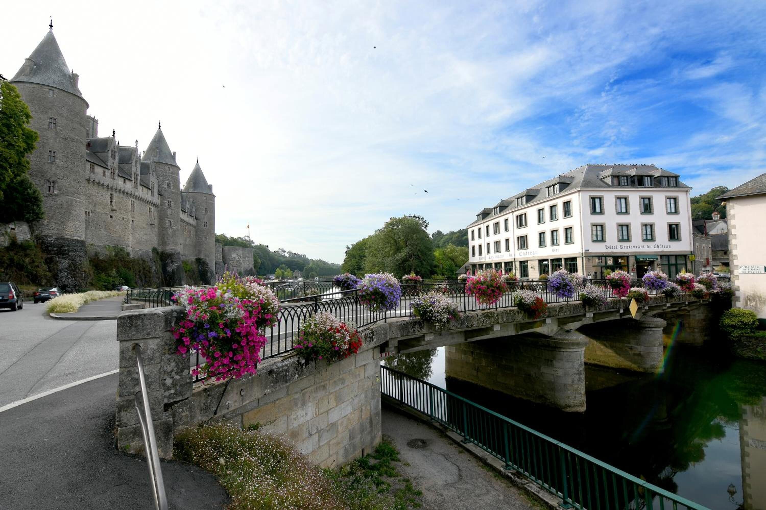 Un château historique en pierre, doté de nombreuses tours, un pont agrémenté de jardinières et un bâtiment moderne avec une enseigne indiquant la présence d’un restaurant.