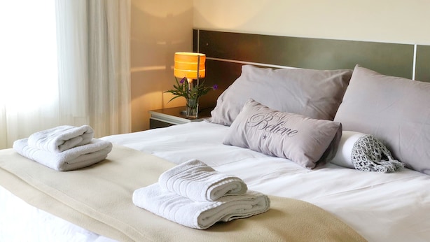 A neatly made bed with folded towels, a bedside lamp, and a decorative pillow with the word "Belair".