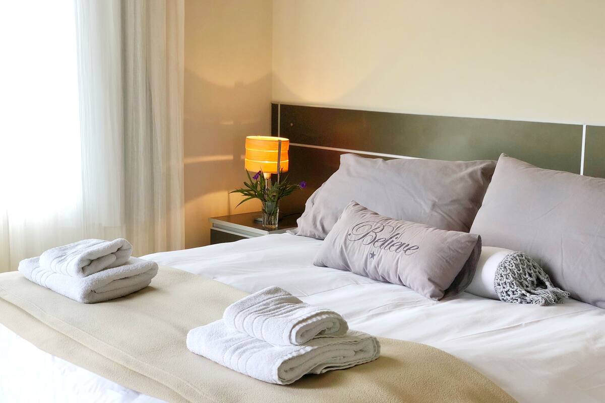 Cama bem arrumada com toalhas dobradas, abajur e travesseiro decorativo com a palavra "Belair".