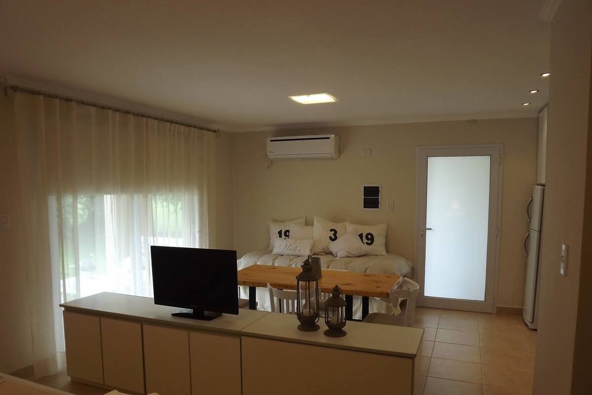 Quarto de hotel com televisão sobre um console de madeira, cama com roupa de cama branca e almofadas numeradas, unidade de ar condicionado e porta que dá para outro quarto.