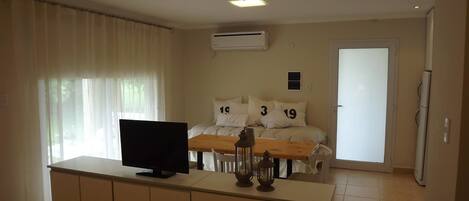 Habitación de hotel con una televisión sobre una consola de madera, cama con ropa blanca y almohadas numeradas, un equipo de aire acondicionado y una puerta que da a otra habitación.