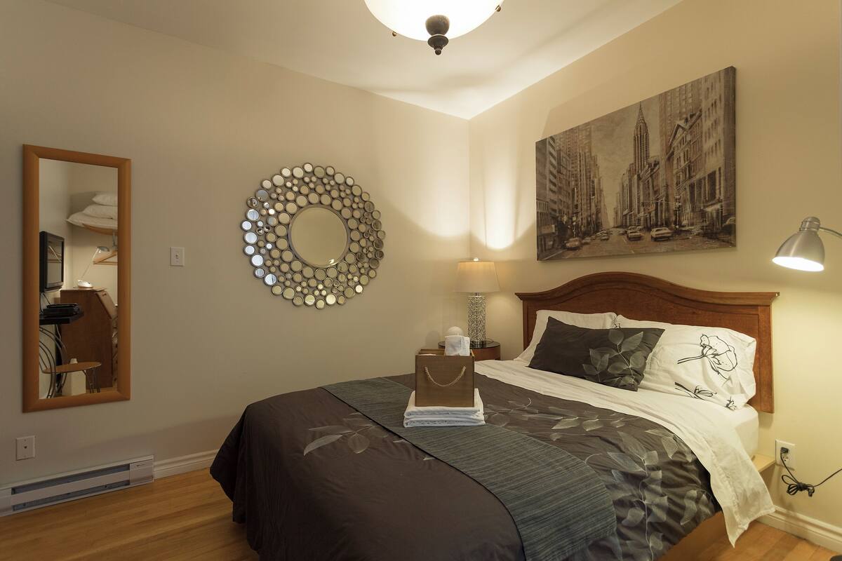 A neatly made bed with a dark bedspread, a headboard with a light wood finish, a round mirror with a circular frame, a wall-mounted light fixture, and a framed cityscape painting.