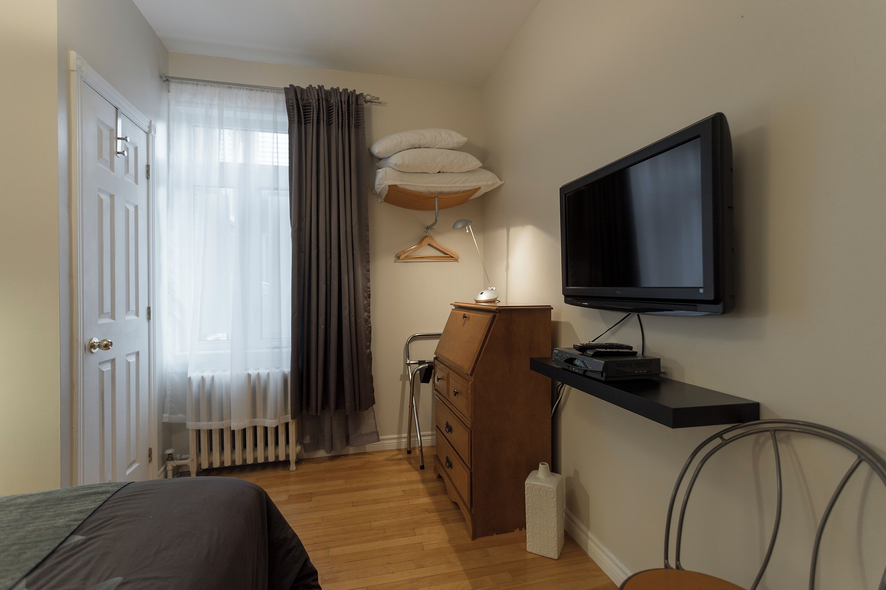 Une chambre à coucher avec un lit, une commode, un téléviseur, un radiateur et une fenêtre avec des rideaux.