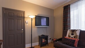 A living room with a dark wood floor, a television mounted on the wall, a small wood-burning stove, a sofa with a blanket and pillow, and a door with a handle.