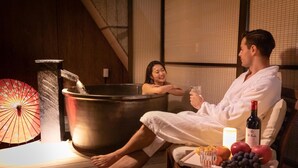 A couple relaxing in a hot tub with a wooden floor and a tiled wall. There is a bottle of wine, a glass, and a tray with fruits on the side.