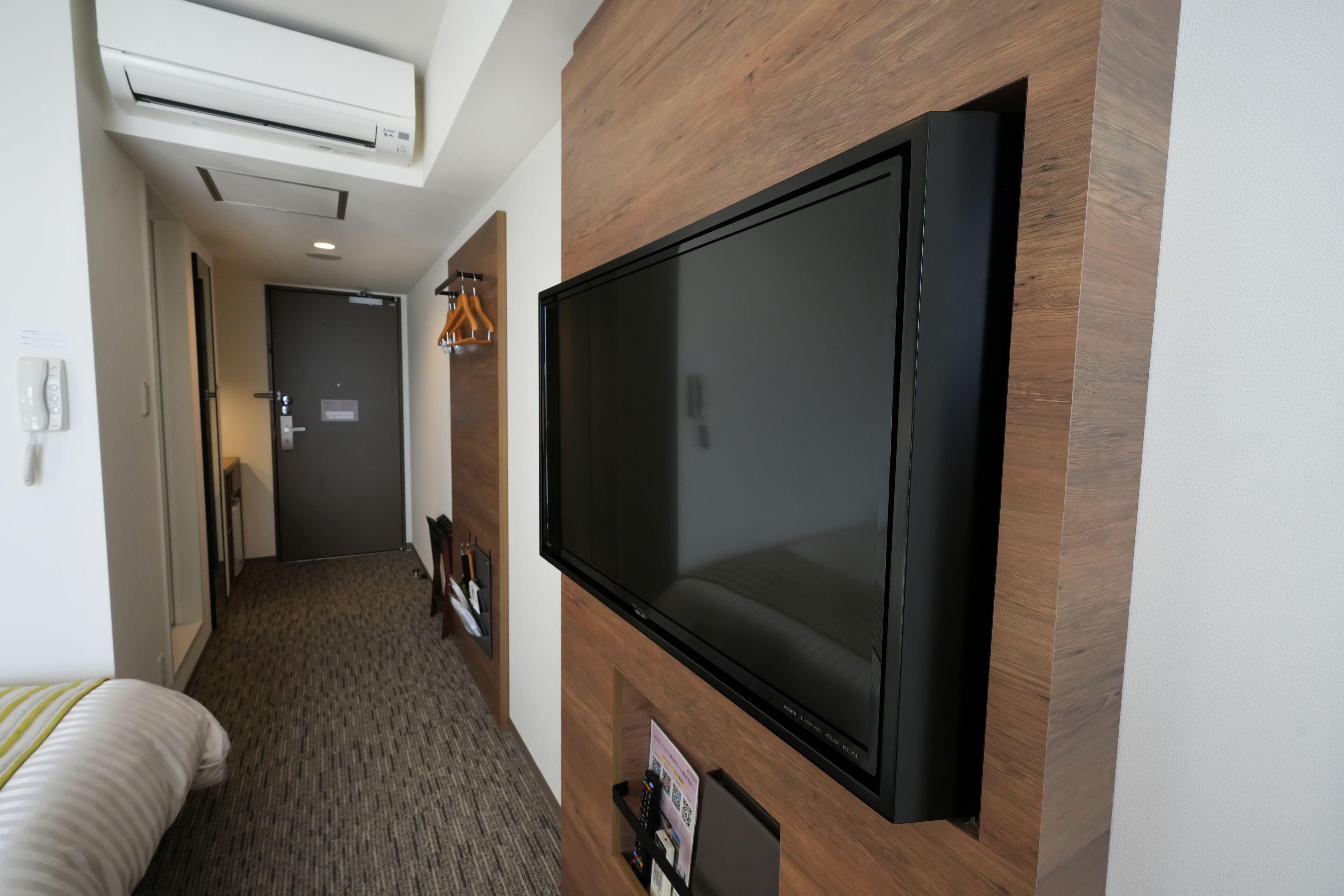 A hotel room with a large flat-screen TV mounted on the wall, a bed with a striped pillow, a carpeted floor, and a wooden accent wall.