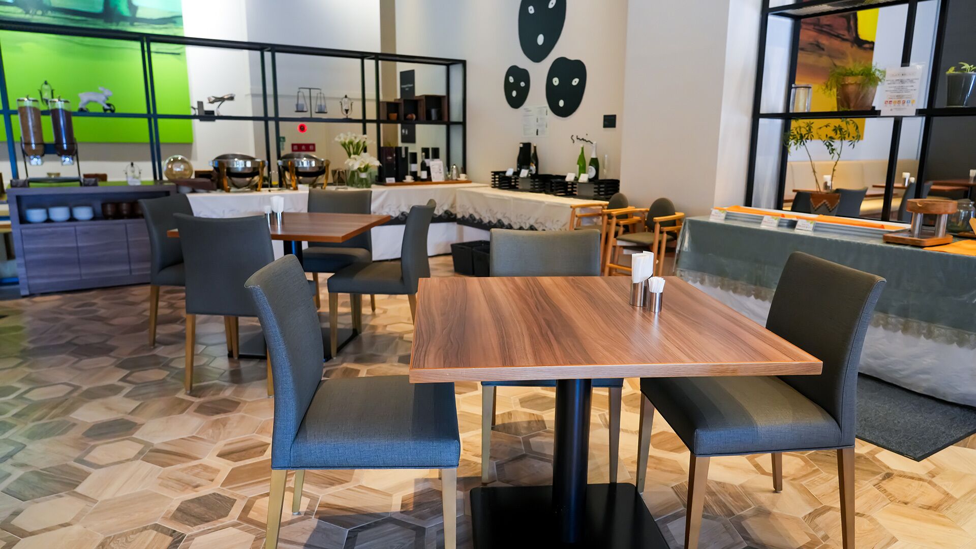 Et moderne restaurantinteriør med hexagonmønstret gulv, træborde, grå polstrede stole og en grøn væg med dekorative genstande.