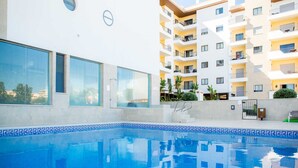 Una piscina con borde de azulejos, junto a un edificio de varios pisos con balcones y ventanas grandes.