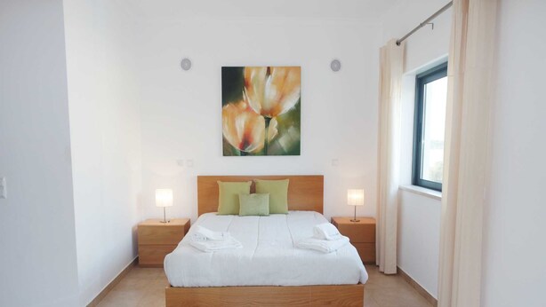 Un dormitorio ordenado con una cama de madera, dos mesitas de noche con lámparas, un ventanal con cortinas y un cuadro de flores en la pared.