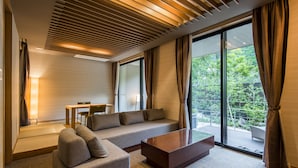 A modern hotel room with a sofa, a coffee table, a dining table with chairs, and large windows offering a view of greenery outside.