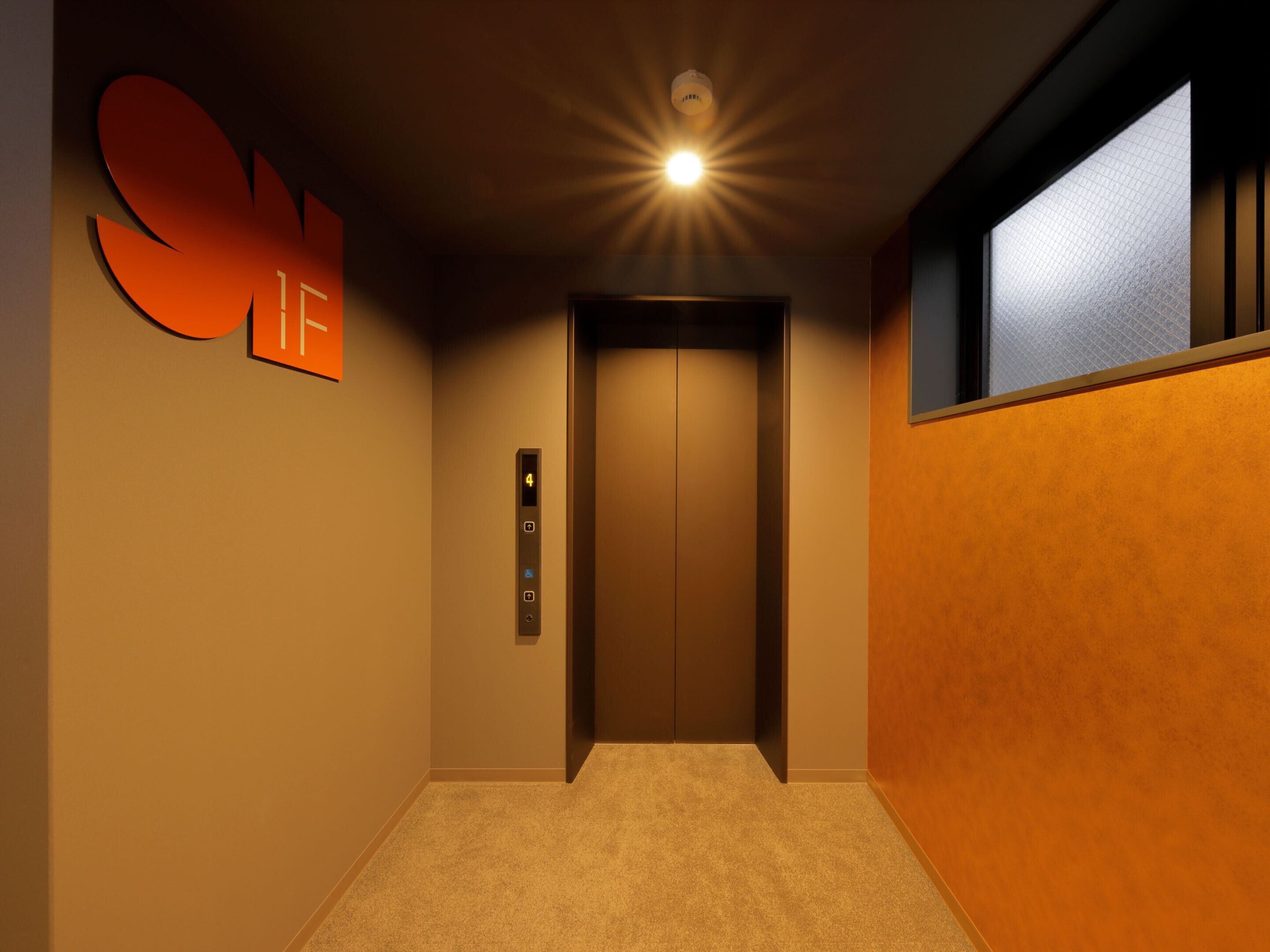 A hallway with a carpeted floor, a wall-mounted elevator control panel, and a large orange sign.