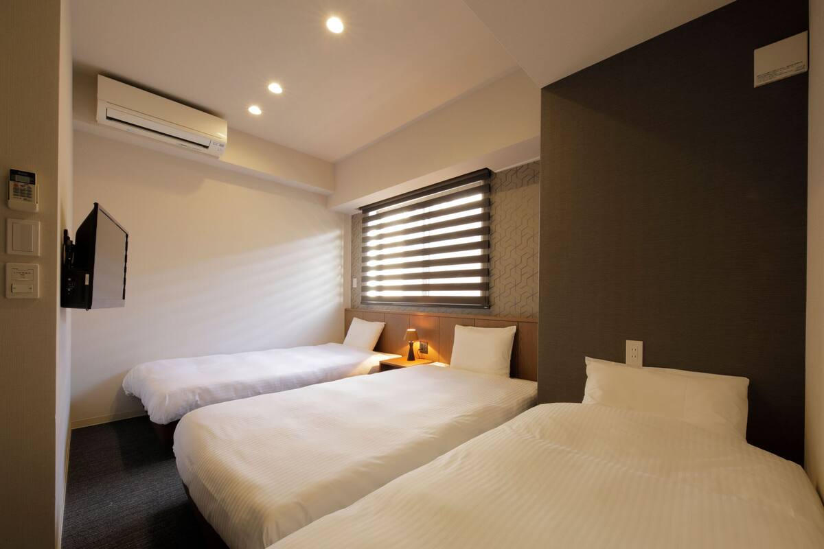 A hotel room with two single beds, an air conditioning unit, a bedside table with a lamp, and a window with blinds.
