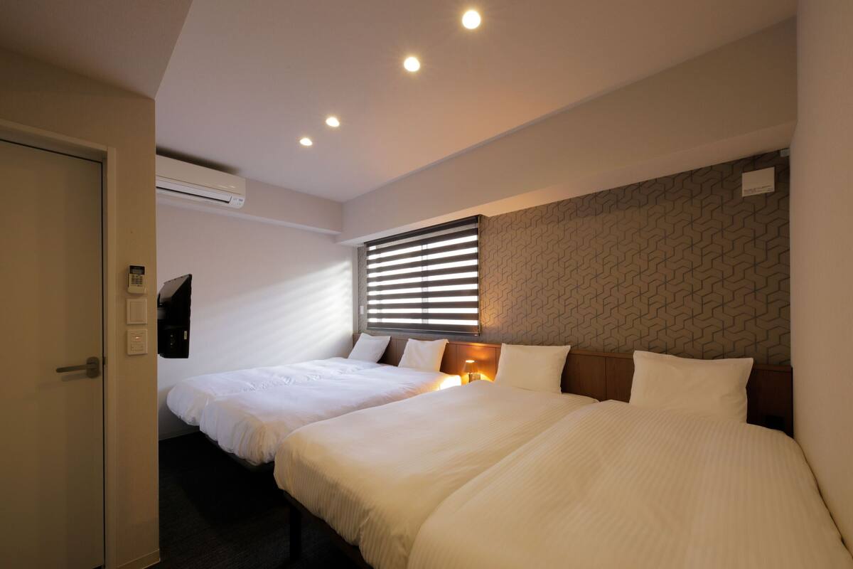 A hotel room with two beds, a wall-mounted air conditioner, a window with blinds, and a patterned wall.