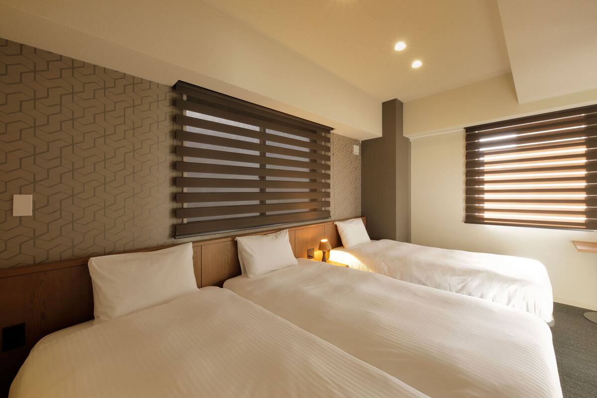 A hotel room with two beds, a wooden headboard, a patterned wall, and a window with blinds.