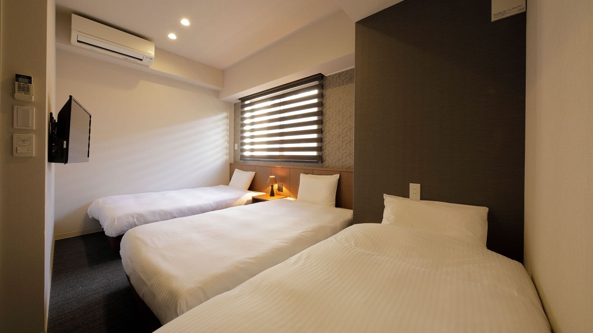 A hotel room with two beds, an air conditioning unit, a wall-mounted phone, and a window with blinds.