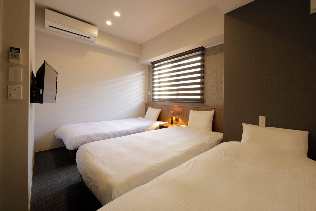 A hotel room with two beds, an air conditioning unit, a wall-mounted phone, and a window with blinds.