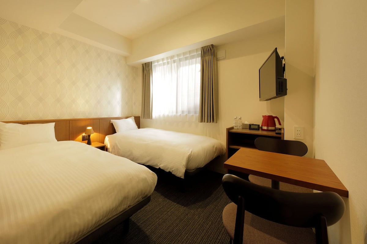 Habitación de hotel con dos camas, un escritorio, una silla, un televisor y una ventana con cortinas.