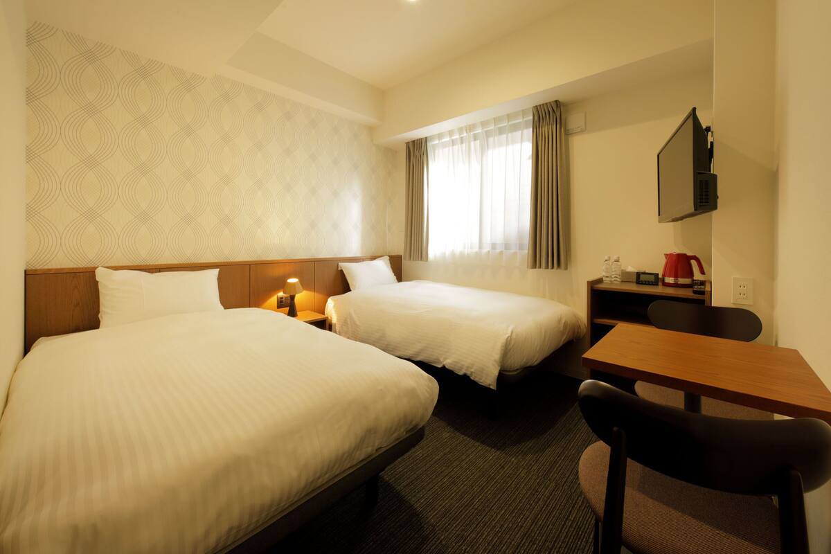 A hotel room with two beds, a desk, a chair, a TV, and a window with curtains.