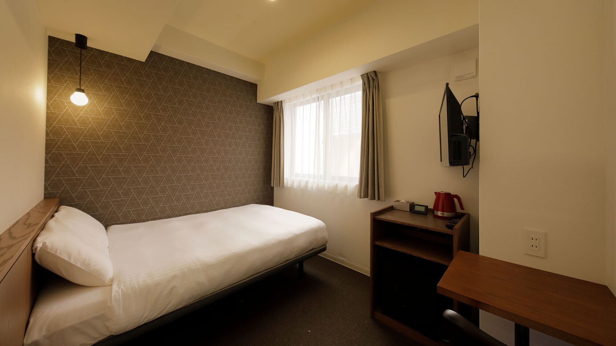 A small, single-bed room with a sloped ceiling, a wall-mounted TV, a bedside table with a kettle, and a window with curtains.