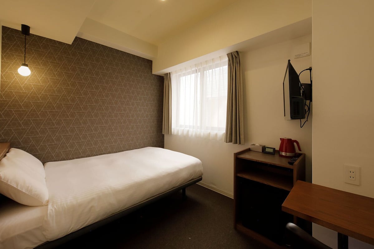 A small, single-bed room with a sloped ceiling, a wall-mounted TV, a bedside table with a kettle, and a window with curtains.