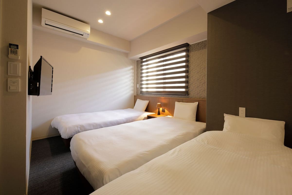 Habitación de hotel con dos camas, un equipo de aire acondicionado, un panel de control en la pared y una ventana con persianas.