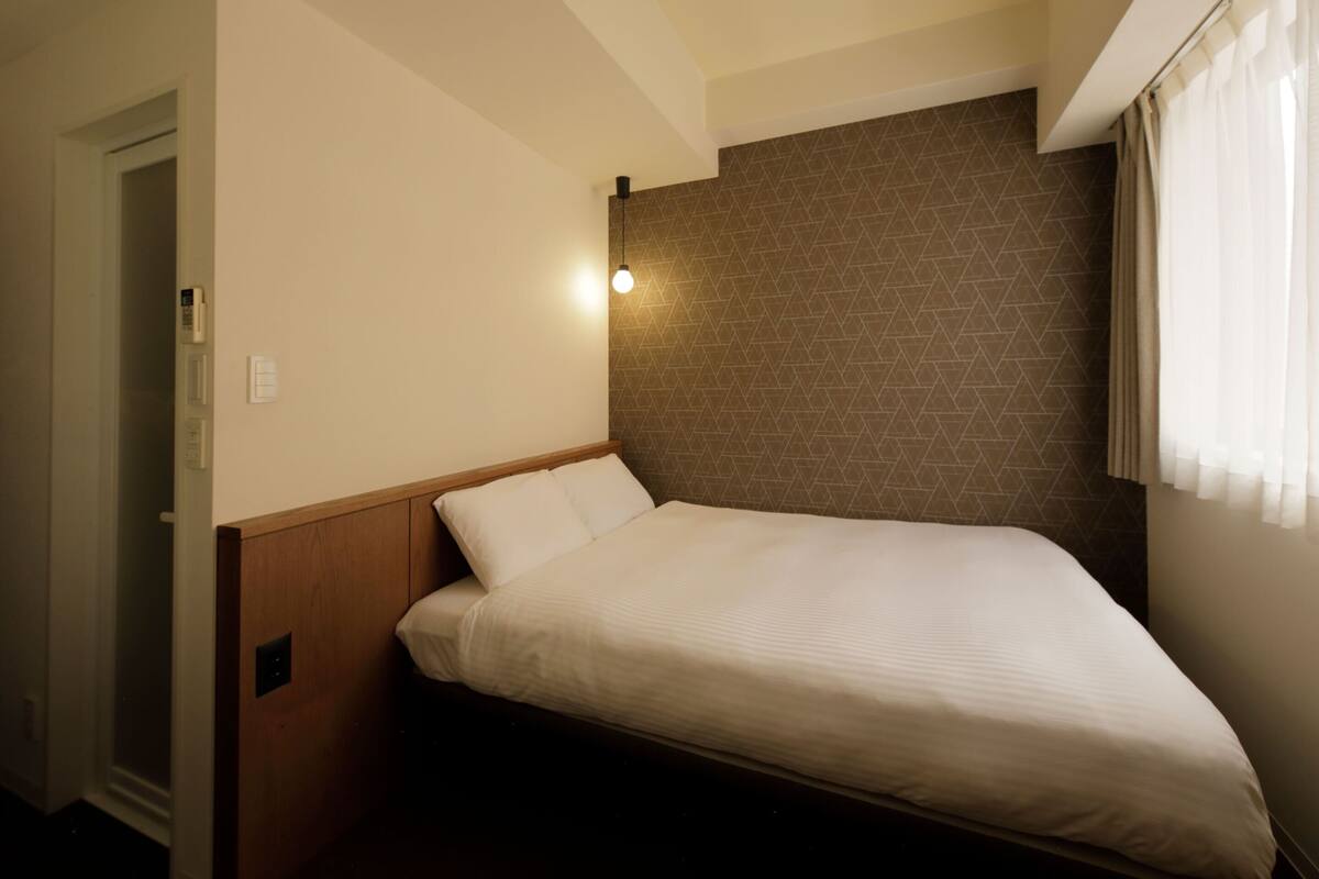 A small, single-bed room with a wooden headboard, a geometric-patterned wall, and a window with curtains.