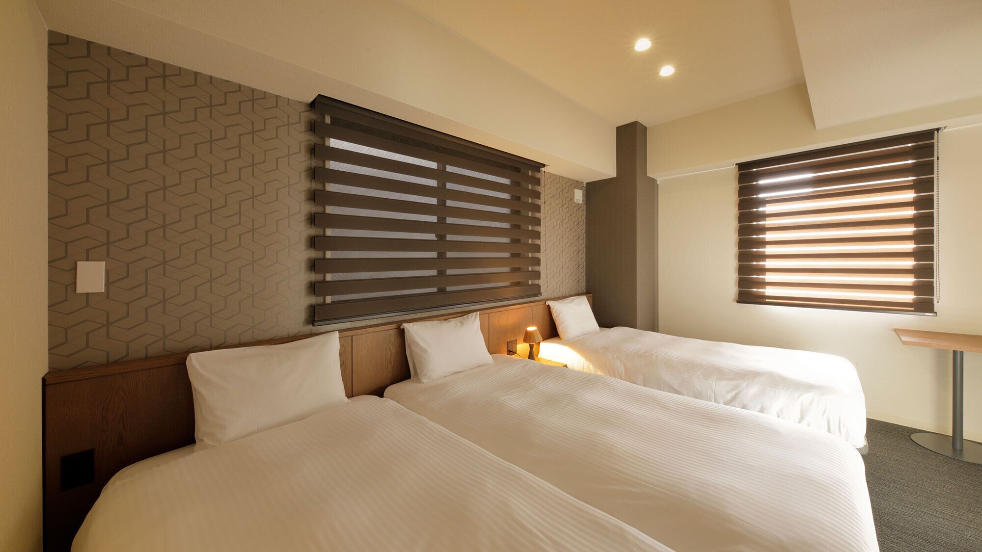 A hotel room with two beds, a patterned wall, and two windows with blinds.