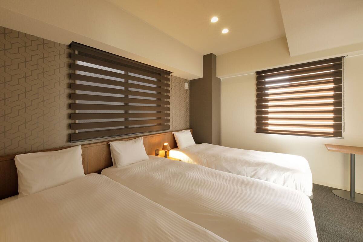 A hotel room with two beds, a patterned wall, and two windows with blinds.