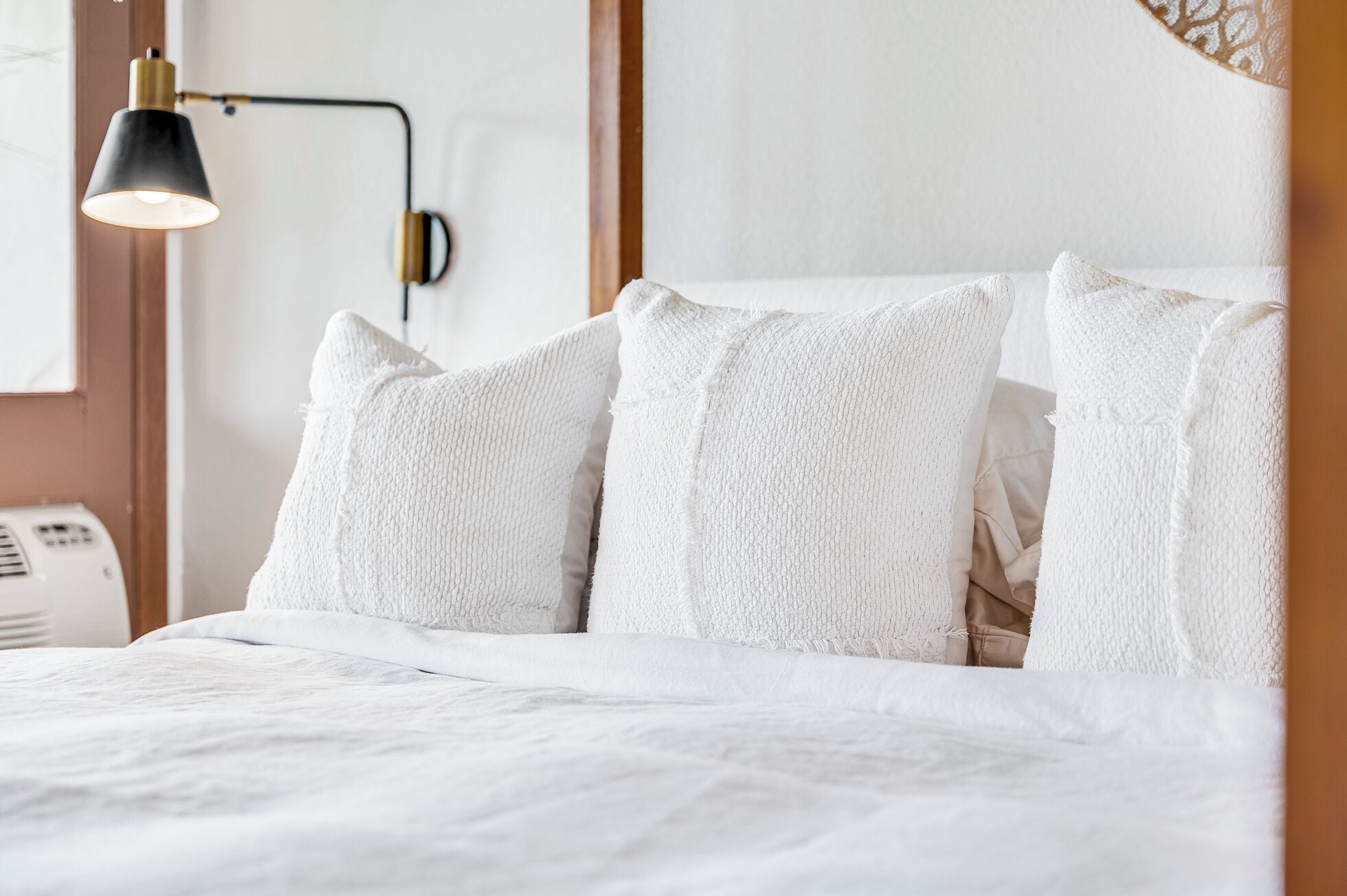 Ein ordentlich bezogenes Bett mit weißer Bettwäsche und Kissen, eine schwarz-goldene Wandlampe und eine Wand mit strukturierter Oberfläche.
