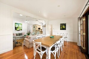 A spacious dining area with a wooden table and white chairs, a kitchen in the background, and a large window with curtains.