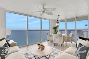 A spacious balcony with a view of the water, furnished with a round table, chairs, and a ceiling fan. - ROMANTIC 5TH-FLOOR BAYFRONT RETREAT | DOLPHINS, SUNRISE VIEWS, POOL, HOT TUB, BEACH GEAR! (Fort Myers Beach)