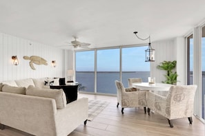 A spacious living room with a large window overlooking the sea, a ceiling fan, and a dining area with a round table.